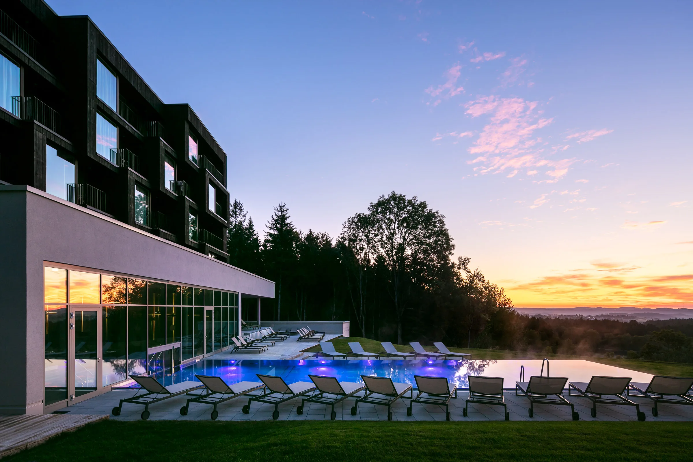 Modern building with a glass facade and illuminated pool at sunset, surrounded by trees and lounge chairs.
